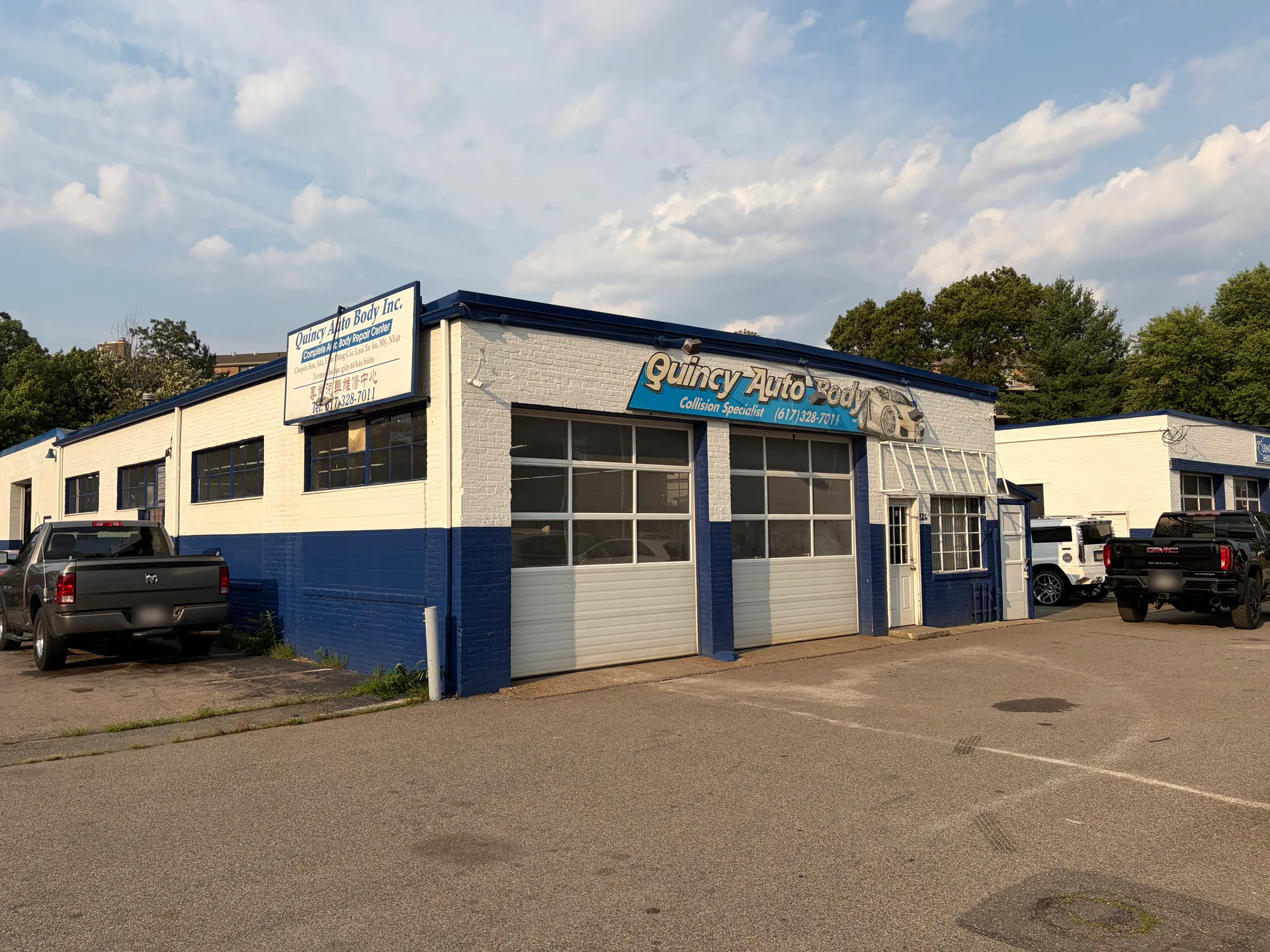 The exterior of Quincy Auto Body on Quincy Ave, with its blue-and-white building, signage, and roll-up bay doors on a sunny day